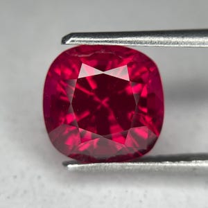 Può includere: Una gemma di rubino sfaccettata a taglio cuscino, tenuta da una pinzetta. Il rubino è di un colore rosso intenso e saturo con riflessi interni. Le sfaccettature della gemma creano un motivo di luce a stella.