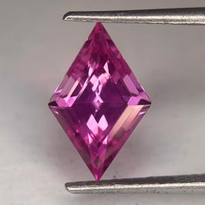 May include: A square-cut, pink gemstone held by tweezers. The gemstone's facets reflect light, highlighting its internal structure. The color is a vibrant pink. The gemstone is likely a precious or semi-precious stone.