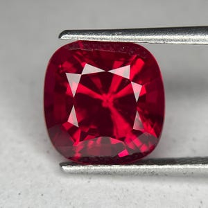 May include: A cushion-cut, deep red gemstone held by metal tweezers. The gem's facets reflect light, creating a brilliant display. The stone's shape is a rounded square, showcasing its rich color and clarity.