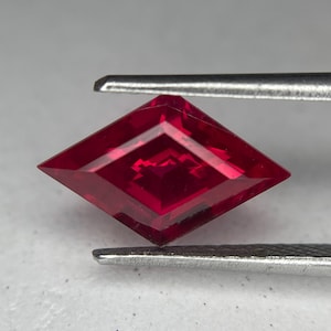 Può includere: Una gemma rosso intenso sfaccettata, tenuta da una pinzetta d'argento. La gemma è tagliata a forma di aquilone, con molteplici sfaccettature che riflettono la luce. Il colore è un rosso ricco e saturo, e la gemma sembra impeccabile.