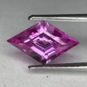 May include: A faceted, kite-shaped gemstone in shades of pink and magenta is held by a pair of tweezers. The gem has a complex internal structure, reflecting light. The gem is a vibrant, eye-catching piece.