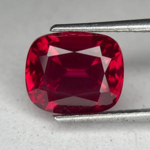 May include: A faceted, cushion-cut ruby gemstone. The gem is a deep, saturated red color, with multiple facets that reflect light. The ruby is held by a pair of silver tweezers, showcasing its shape and brilliance. This gemstone is ideal for jewelry.
