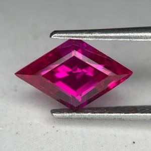 May include: A faceted, kite-shaped gemstone with a deep magenta hue. The gem is held by silver tweezers, showcasing its sharp angles and internal facets. The gemstone's color is a vibrant, saturated pink, reflecting light.