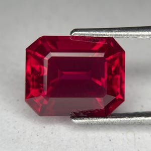 May include: A faceted, rectangular ruby gemstone held by a pair of silver tweezers. The ruby is a deep, saturated red color with sharp, clean edges and internal facets that reflect light. The gem is cut in an emerald shape.