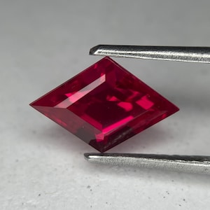 以下が含まれることがあります： 銀色のピンセットで保持された、ファセットカットされた深紅色の宝石。 宝石は鋭角と光を反射する研磨された表面を持つ凧型です。 宝石の色は豊かで飽和した赤色で、ルビーのように見えます。