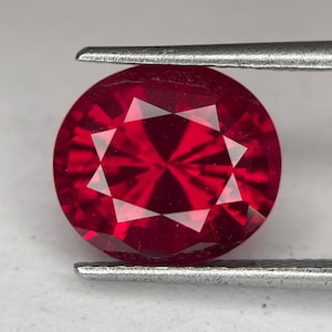 May include: A faceted, round-cut ruby gemstone held by tweezers. The gemstone is a deep, vibrant red color with multiple facets that reflect light. The tweezers are silver-colored and hold the gemstone securely.