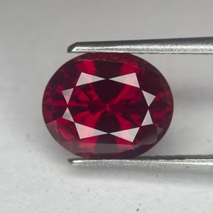 May include: A faceted, oval-cut, deep red gemstone. The gem is held by silver tweezers, showcasing its intricate internal facets. The stone's color is a rich, saturated red, with a brilliant, reflective surface. This gemstone is likely a ruby.