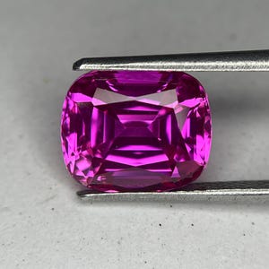 May include: A vibrant, cushion-cut gemstone in a deep magenta hue. The gem is held by a pair of metal tweezers, showcasing its facets and internal structure. The stone's color is consistent throughout, with a high degree of clarity.