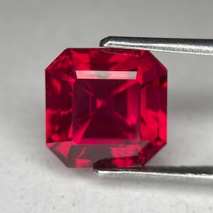 May include: A square-cut, deep red gemstone, held by silver tweezers. The facets of the gem are precisely cut, reflecting light to create a brilliant display. The gemstone's rich colour and clarity are highlighted by the tweezers.