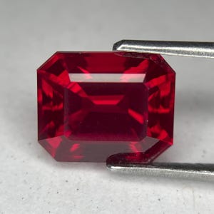 Könnte beinhalten: Ein facettierter, rechteckig geschliffener Rubin-Edelstein. Der Edelstein hat eine tiefe, leuchtend rote Farbe und wird von einer Pinzette gehalten. Der Schliff des Steins erzeugt mehrere Facetten, die das Licht reflektieren.