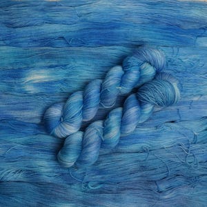 Puede incluir: Primer plano de madejas de hilo azul y hebras sueltas. El hilo es una mezcla de azules claros y oscuros, creando una apariencia texturizada similar al océano. Las madejas están retorcidas y dispuestas sobre el hilo suelto, mostrando la textura y las variaciones de color del producto.