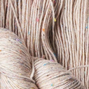Può includere: Primo piano di filato beige con macchie colorate e fili dorati. Il filato è strettamente avvolto in matassa e appare anche come fili sciolti. Questo filato è adatto per progetti di maglieria e uncinetto.