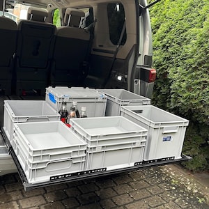 Könnte beinhalten: Ein silberner Minivan mit offener Heckklappe, der ein ausziehbares Schubladensystem mit fünf grauen Plastik-Aufbewahrungsbehältern zeigt. Die Behälter sind mit verschiedenen Gegenständen gefüllt, darunter Getränkeflaschen.