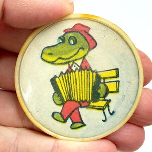 May include: A vintage, round button featuring a cartoon alligator playing an accordion while seated on a yellow bench. The alligator wears a red hat, jacket, and pants. The button has a yellowed, aged appearance.