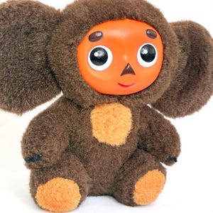 Op de afbeelding: Een pluche speelgoed met een bruin, harig lichaam en grote, ronde oren. Het heeft een oranje gezicht met grote ogen met zwarte pupillen, een kleine neus en een lachende mond. De borst en voeten hebben oranje vlekken.