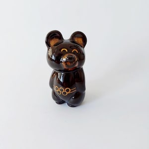 May include: A glossy dark brown ceramic bear figurine featuring a rounded head, small ears, and a smiling face. The bear has a gold-coloured Olympic rings emblem printed on its chest, with a smooth, reflective finish across the entire surface.