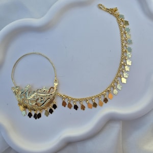 Op de afbeelding: Een goudkleurige neusring met een cirkelvormige ring verbonden aan een decoratief stuk met geëtste bladachtige patronen. Een fijne ketting strekt zich uit vanaf de zijkant, versierd met kleine, bungelende bladvormige bedeltjes.