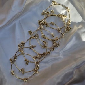 Puede incluir: Brazaletes dorados con racimos decorativos de pequeñas cuentas. Los brazaletes están colocados sobre una tela blanca y sedosa, creando una exhibición suave y elegante. La luz se refleja en los brazaletes y la tela.