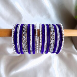 Puede incluir: Un conjunto de elegantes pulseras en tonos morado, blanco y plateado. Las pulseras están dispuestas sobre una varilla de madera sobre un fondo de tela blanca. El diseño incluye elementos de terciopelo, abalorios y joyas.