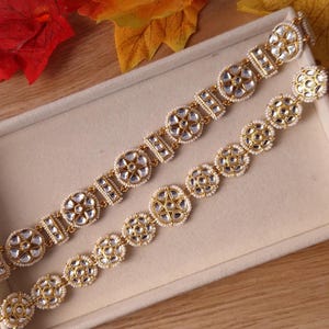 May include: Two gold-toned bracelets with intricate designs. Each bracelet features circular and geometric patterns with clear, reflective accents and small, white beads. The bracelets are displayed in a light-colored box, with autumn leaves in the background.