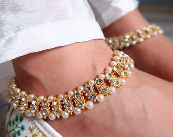 Könnte beinhalten: Goldfarbene Fußkettchen mit Kunstperlen und klaren Kristallen. Die Fußkettchen werden über weißen Schuhen mit farbenfrohen Blumenstickereien getragen. Das Bild zeigt auch eine weiße Hose und den Text "KAURKHAZANA".