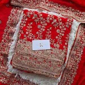 May include: A red embroidered textile set with gold floral patterns. The set includes a top, bottom, and scarf, all adorned with intricate gold embroidery. A small white card with text is placed on the top. The set is displayed on a white fluffy surface.