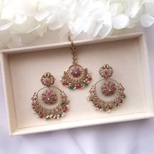 May include: A three-piece jewelry set featuring a gold-toned tikka and matching earrings. The earrings and tikka have a circular design with red, green, and white accents. The set is displayed in a white box.