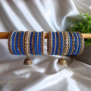 Puede incluir: Un conjunto de pulseras azules y doradas con detalles intrincados. Las pulseras están adornadas con pequeñas cuentas doradas, perlas blancas y adornos dorados. Cada conjunto incluye un dije en forma de campana dorada. Las pulseras se exhiben sobre una varilla de madera sobre un fondo de satén blanco.