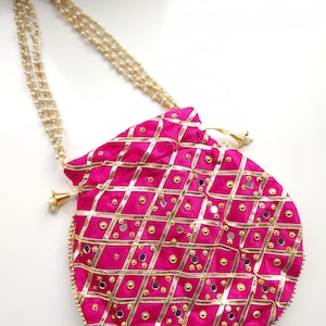 Könnte beinhalten: Eine pinkfarbene runde Beuteltasche mit einem goldperlenbesetzten Riemen. Die Tasche hat ein goldfarbenes Gittermuster mit kleinen Spiegeln und goldenen Verzierungen. Der Kordelzugverschluss ist mit einer goldenen Quaste versehen.