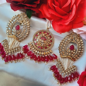 May include: A set of three gold-toned jewellery pieces with pink and white accents. The pieces include a pendant, a pair of earrings, and a maang tikka. The jewellery is decorated with intricate designs and features a combination of pink and white beads.