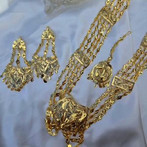 May include: A gold-toned jewelry set consisting of a multi-strand necklace with a large central pendant, a pair of dangling earrings, and a matching headpiece. Each piece features intricate etched floral patterns and delicate chain links.