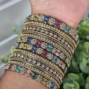 May include: A stack of gold-toned metal bangles featuring embedded clear rhinestones and small, oval-shaped multicoloured stones in shades of pink, blue, green, and yellow. The bracelets have a textured, antique-style finish and are worn together.