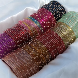 May include: A selection of vibrant glass bangles in various colours, including red, pink, green, and brown, each decorated with tiny gold-toned details. The bangles are arranged in a white, satin-lined container, offering a striking visual presentation.
