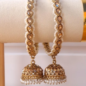 May include: A pair of gold-toned bangles featuring a row of teardrop-shaped champagne-coloured stones bordered by small white pearls. Each bangle is attached to a bell-shaped jhumka earring adorned with matching stones and a fringe of tiny white pearls.