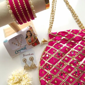 Könnte beinhalten: Eine Sammlung indischer Schmuckstücke und Accessoires, darunter ein pinkfarbenes Samtbanglen-Set mit Perlenakzenten, eine pinkfarbene Handtasche mit goldenen Verzierungen und Spiegeldetails sowie goldene Jhumka-Ohrringe. Eine weiße Blume und Kristallverzierungen sind ebenfalls vorhanden.