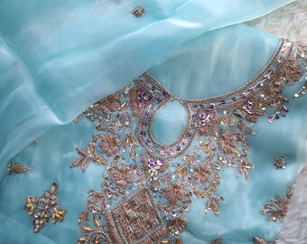 May include: A light blue, sheer fabric garment with intricate embroidery. The embroidery features gold and bronze thread, sequins, and beadwork. The neckline is embellished with a detailed pattern. The garment is likely a formal dress or top.