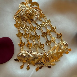 Op de afbeelding: Een goudkleurige decoratieve broche met een centraal bloemachtig motief met textuur en geëtste lijnen. Vijf delicate kettinkjes hangen aan het bovenstuk, elk versierd met kleine bladvormige bedels, eindigend in een bredere, gebogen basis met bladpatronen.