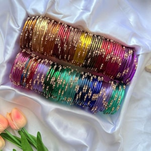 May include: Two sets of colourful bangles, each with multiple glass bracelets in shades of gold, red, pink, green, blue, and purple. The bangles are decorated with small, gold-coloured embellishments, displayed on a white satin surface.