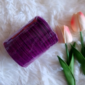 May include: A bundle of deep purple velvet ribbons, wrapped in clear plastic, rests on a fluffy white surface. Artificial tulips with pink and white petals and green stems are in the background. The ribbons appear to be new and unused.