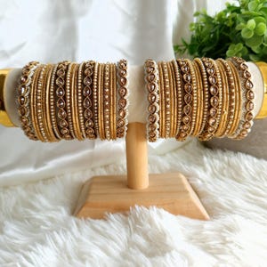 May include: Two sets of gold bangles displayed on a wooden stand. Each set features multiple bangles with varying designs, including beaded and gem-studded accents. The bangles are arranged on a white, fluffy surface.