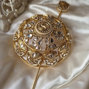 Pode incluir: Um acessório de cabelo em metal dourado com uma estrutura em forma de cúpula adornada com pedras transparentes em forma de gota e contas brancas. O centro apresenta um motivo circular com uma figura de pássaro e um alfinete dourado fino.