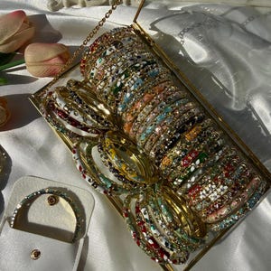 May include: A selection of colourful bangles in a gold-framed glass box. The bangles have various designs with red, white, blue, and green accents. A small, matching bangle is displayed in a white pouch.