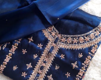 May include: A navy blue embroidered outfit with a matching sheer scarf. The outfit features intricate gold and pink floral embroidery along the neckline and bodice. The fabric has a subtle sheen, and the scarf drapes elegantly over the ensemble.