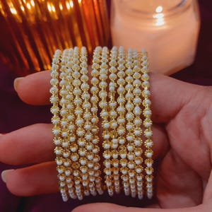 May include: A stack of gold bangles with white beads. The bangles are stacked on top of each other and are all the same size. The beads are small and round and are evenly spaced around the bangles.