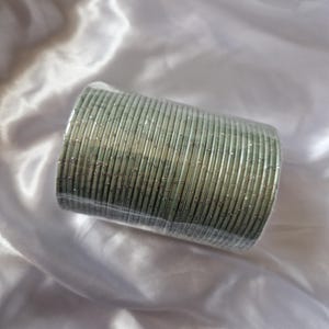 Puede incluir: Un conjunto de pulseras verde plateado envueltas en plástico transparente. Las pulseras están apiladas, formando un cilindro. El fondo es una tela blanca y sedosa.