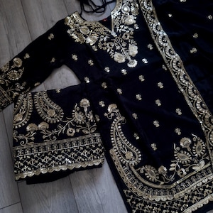 May include: A black velvet outfit with detailed gold embroidery. The set comprises a long-sleeved top and a matching scarf. The gold embroidery showcases floral and paisley designs, providing an elegant aesthetic.