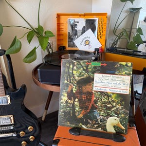 Può includere: Un amplificatore arancione con un disco in vinile vintage sopra. La copertina del disco presenta un bambino con una camicia a quadri e un'anatra. Il disco è intitolato "Pierino e il lupo" e "Lo schiaccianoci". Una chitarra elettrica nera è visibile a sinistra.