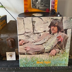 May include: A vintage vinyl record album cover featuring James Taylor. The cover art shows James Taylor lying on a grassy patch in front of a stone wall. He is wearing a brown suit with a patterned tie and a white shirt. The album title "James Taylor" is printed in yellow at the bottom of the cover.