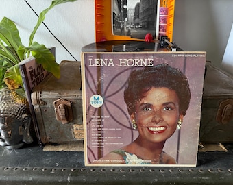 Pode incluir: Capa de álbum de vinil vintage com Lena Horne. A capa apresenta um retrato de Horne com uma assinatura. O álbum é rotulado "TOPS" e inclui títulos de músicas. O disco está em uma vitrola.