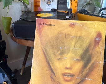 May include: A square vinyl record sleeve for The Rolling Stones featuring a blurred, golden-toned portrait of a face. The album cover is positioned in front of a black turntable with a yellow record label and a white Deadwax Goods sticker.